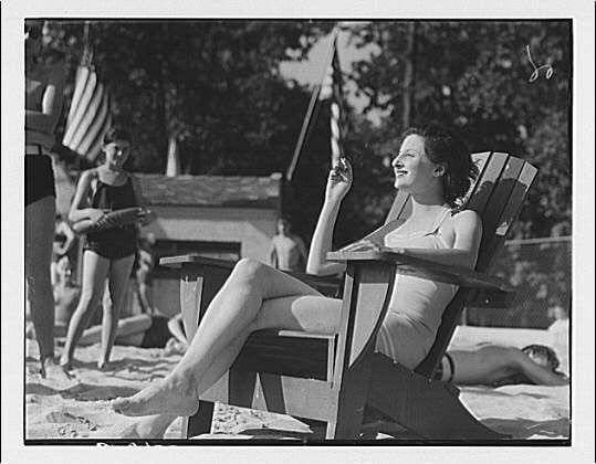 The black and white photograph captures a woman lounging on an Adirondack chair at what appears to be Glen Echo amusement park. She is dressed in a light-colored swimsuit, with her right arm raised holding onto the backrest of the chair as if she's shielding her eyes from the sun or enjoying a bright day outdoors. Her left leg extends outward casually over one side of the wooden seat.

In the background, there are several individuals engaging in various activities typical of a beach setting: swimming and lounging on towels by what could be an outdoor pool area adorned with American flags. The overall scene suggests leisurely summer days spent enjoying water-related recreation at this amusement park destination popular during its time period.