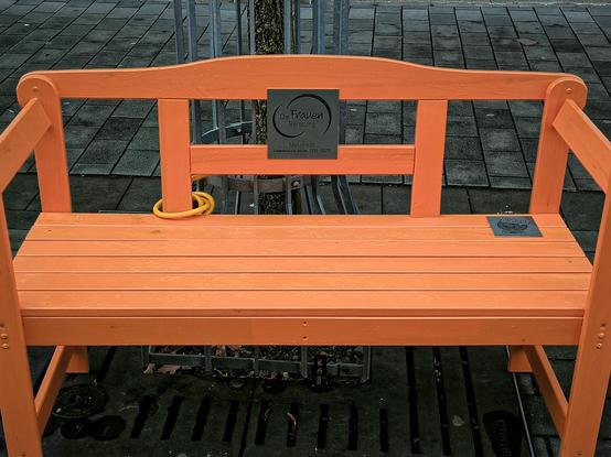 Orange Bank von schräg oben fotografiert. Zwei Plaketten. Eine an der Rücklehne, die andere auf der Sitzfläche rechts. Text siehe Beitrag.