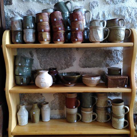 Un petit meuble en bois de 3 étagères où sont empilées plein de pièces de céramique. On y trouve des tasses à café, des mugs, des bols, des vases, des bouteilles et des boîtes. Les couleurs dominantes sont le blancs, le rouge et le vert.