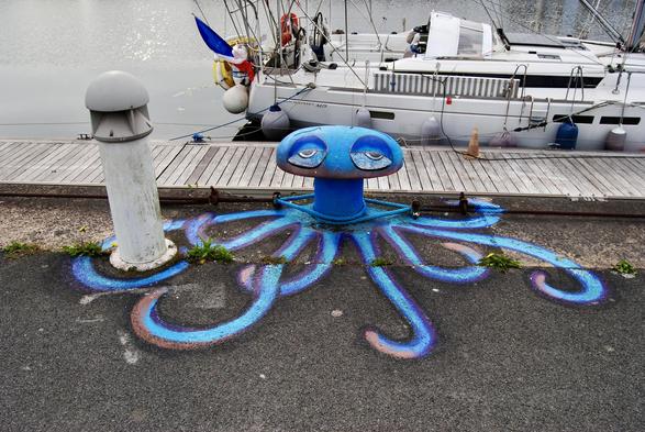 Künstlerische Darstellung eines blauen Oktopus in Verbindung mit einem Poller für Boote, welcher den Kopf mit müden Augen darstellt. Boote sind im Hintergrund zu sehen, die an einem Yachthafen angedockt sind.