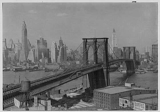 The image depicts a black and white photograph of the Brooklyn Bridge spanning across the East River in New York City. In the foreground, we can see the bridge's structure with its distinctive suspension towers supporting cables that hold up the roadway above it. The Manhattan skyline is visible on the left side of the photo, showcasing iconic skyscrapers like the Empire State Building and One Chase Manhattan Plaza.

The Brooklyn Bridge itself appears in two segments: a closer view crossing over the East River, and another segment extending into the distance toward Downtown Brooklyn. In the background, we can see other bridges connected to Long Island City on the opposite shore of the river. The overall composition captures an industrial atmosphere with warehouses along the waterfront and signage for businesses such as "COFFEE" indicating commercial activity in this urban environment.

The photograph is labeled as a negative from Samuel H. Gottscho's collection, taken at Robert Gair Building on April 1st, 1932, providing historical context to its creation during that period of the Great Depression and early World War II era.