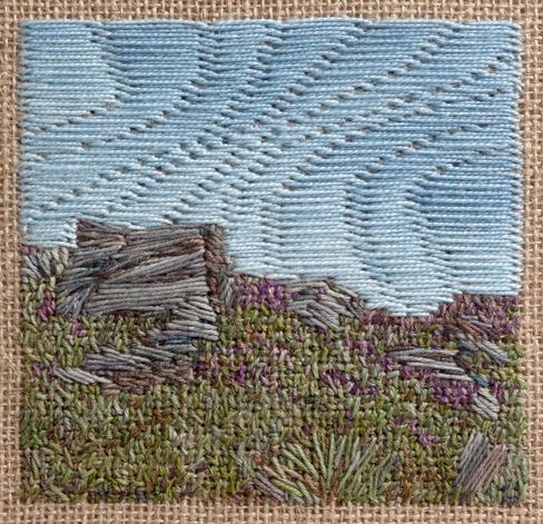 Stitched artwork depicting a heather and grass covered bank with a large rock and part of a dry stone wall to the left. A few scattered rocks to the right. The sky is clear and blue.