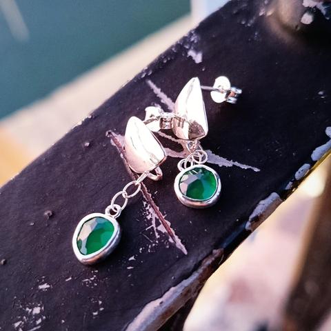 a pair of silver earrings with a green gemstone each. they have a silver triangle and from there is connected with a small silver chain the gemstone. they are placed on an old black handrail