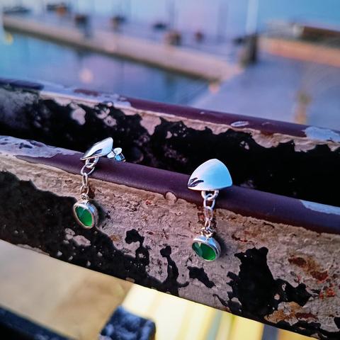 a pair of silver earrings with a green gemstone each. they have a silver triangle and from there is connected with a small silver chain the gemstone. they are placed on an old black handrail