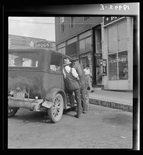 The black and white photo shows two men beside a vintage automobile, which appears to be parked on the street. One man is leaning against the back of an older car with its gas tank attached; he wears overalls, a long-sleeved shirt under his rolled-up sleeves, suspenders, boots, and a hat, suggesting work attire or manual labor. The other stands beside him in similar clothing. In front of them are buildings that house businesses: one appears to be a drugstore with "DRUGS" clearly visible on the sign above its entrance; another has signs indicating it is for cigars, meat market goods. There's also an obscured store name starting with 'CORN'. A few pedestrians can be seen in the background, and some items of interest like bottles or containers attached to the back of the car suggest a delivery purpose. The photograph seems candid, capturing everyday life possibly from mid-20th century America as suggested by fashion styles and automobile design.