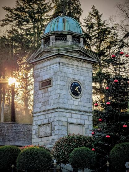 A tall, grey stone clock tower stands prominently against a backdrop of tall trees. The tower features a green, oxidized copper dome and a black clock face with gold hands and numerals. The Roman numerals "MDCCCXLIII" are carved into the stone above the clock. Bright, golden sunlight flares through the trees on the left, creating a warm glow. To the right, a dark pine tree is decorated with large, shiny red Christmas baubles, and manicured green bushes line the bottom of the frame.
