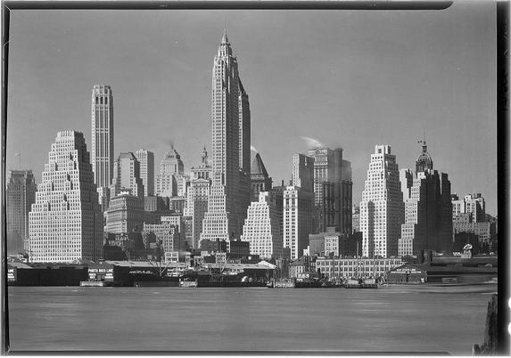 The image depicts a black and white photograph of the skyline of New York City, specifically showcasing numerous tall buildings with classic architecture. The most prominent structure is the Empire State Building in the center background, featuring its iconic spire reaching into the sky. To its left stands another skyscraper with setbacks near the top floors to create small terraces or observation decks.

The photograph captures a cityscape viewed across what appears to be the East River, as evidenced by the boats and barge docked at piers on the waterfront in the foreground. The water reflects some of the urban scene but is mostly calm, suggesting clear weather conditions when this image was captured.

The skyline includes various other buildings with different architectural details such as setbacks, cornices, and a mix of rectangular windows arranged symmetrically or grouped together for aesthetic appeal. Some older structures have more classical features like columns, while others exhibit modernist designs from the early 20th century during which this photo might be taken.

The photograph is noted to come with additional information in "New York city views: Lower New York," dated April 4, 1932, and has been attributed to Samuel H. Gottsch at the Library of Congress collection.