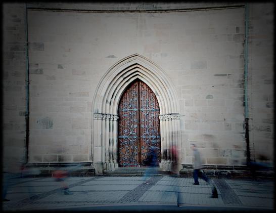 Blick auf ein Kirchenportal. Vor dem Portal sieht man schemenhaft vorbeigehende Menschen (Langzeitbelichtung).