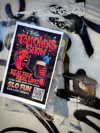 The poster has a black background with red lettering. A man with glasses and a beard is holding a mug of frothy beer and has his mouth open suggesting he's laughing or talking loudly. The text says, "The Tom Owens Show. Real talk. Real life! Every week with Tom Owens. SLO FUN San Luis Obispo." There is a scan QR code for more info. The show poster is taped at a diagonal on a gray utility box that has black graffiti on it.