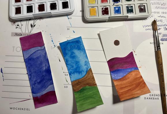Blick auf eine Schreibunterlage. Darauf Aquarellfarben, Pinsel und drei Lesezeichen, die bunt bemalt sind.