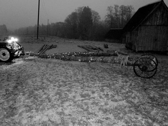 Nightvision shot of the tractor with >5m long log lifted between the tree point hitch and the log wheels on the rear. Another log is confusingly already on the ground behind.