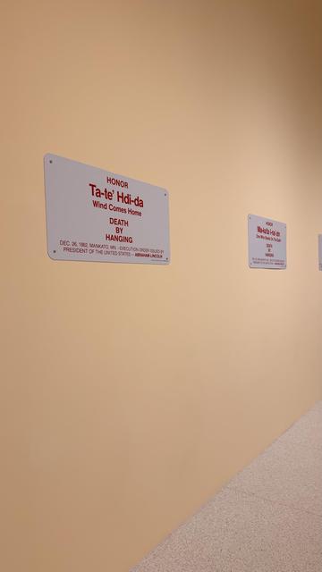 A softly lit beige wall at the Walker Art Center in Minneapolis displays a series of rectangular commemorative plaques. The nearest plaque honors Ta-te’ Hdi-da, “Wind Comes Home,” noting death by hanging on December 26, 1862, under an execution order issued by Abraham Lincoln. Additional similar plaques line the wall in the distance.