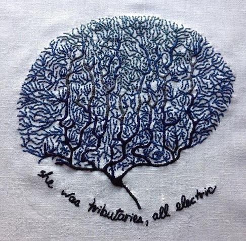 An embroidery on bone white linen. There are many coral-like neurons in the vague shape of a brain embroidered in shades of blue. The bottom section is navy blue and transitions to a lighter blue at the top. The text at the bottom reads: she was tributaries, all electric.