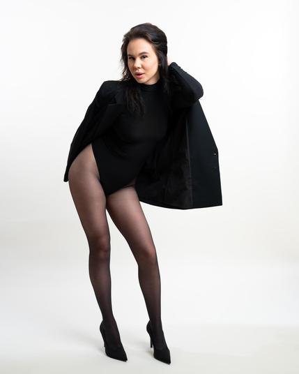 Woman wearing a black leotard, sheer tights, and high heels, with a black jacket draped over her shoulder, standing against a plain white background. Her head is tilted slightly, and one hand holds the jacket while the other rests on her hip.