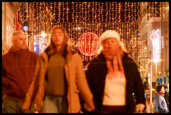 Night-time photograph of pedestrians walking on Oliver Plunkett Street, Cork, beneath cascading golden fairy lights creating a canopy effect overhead, large red neon-illuminated festive spheres and decorative motifs suspended above the street, blurred figures in winter clothing in motion, storefront signage visible at street level.