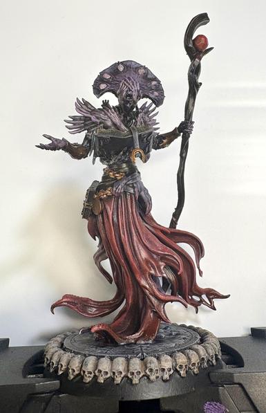 A creepy looking sorcerer holding a staff and a book. It is floating on a dark red and purple cloak and has half a dozen arms or so around its chest. Two bony hands cover its eyes and its skin is a sickly purplish-grey. A circle of skulls surrounds the miniature’s base all the way around.