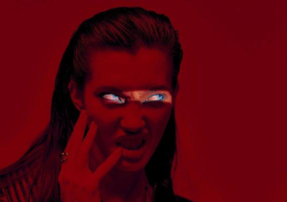 A figure in a black leather jacket illuminated by a light beam across her blue eyes stands against a deep red background, exuding a bold, mysterious vibe.