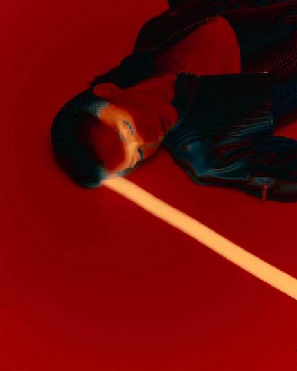 A model in a sleek black jacket lies on a vibrant red background, illuminated by a striking yellow light beam across their forehead.