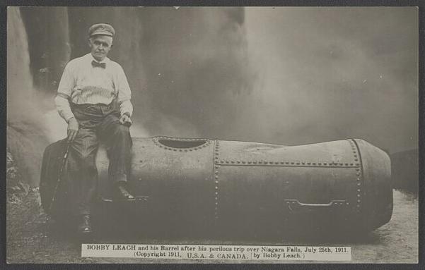 A photograph of a man in a cloth cap, white shirt with bow tie and light colored suspenders, wearing dark trousers and dress shoes, sitting atop a steel tank with riveted sections, a hole in the top, and handles to the side, just a bit larger than the man himself. He is posed on this tank before the mists of Niagara Falls. In his right hand is a cane, presumably necessitated when he broke his knees going over the falls in this barrel.