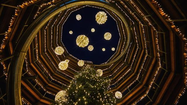 A low-angle photograph looking straight up inside a towering, spiral wooden structure at night. The structure is built in octagonal tiers that narrow towards an open view of the dark blue night sky. Strings of warm, golden lights line the wooden railings of the spiral walkway. Large, glowing spherical ornaments hang suspended in the central void, floating above the illuminated top of a large Christmas tree visible at the bottom of the frame.