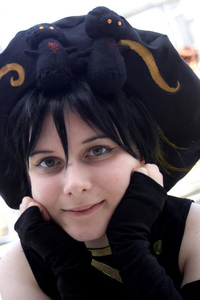 A person with a black poofy hat looking at the camera. On the hat are two black plushie mushroom organization XIII and golden decoration.
The person has short black hair with two yellow curls on their left.