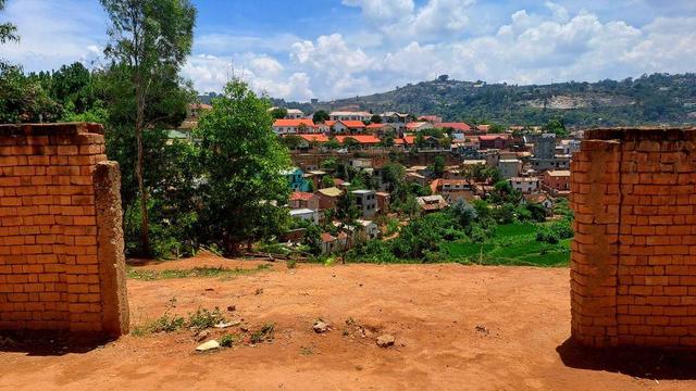 A scenic view through a partially demolished brick wall, revealing a lush, green gully and a hillside neighborhood in Antananarivo, Madagascar. The foreground features two brick pillars framing the view, while the background showcases colorful houses with red and orange rooftops nestled among trees and greenery under a bright, partly cloudy sky.
