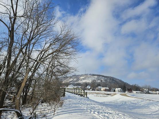 White clouds in a blue sky over a bluff. Leafless trees are next to a path of footprints in white snow, leading to a bridge with brown wooden rails.