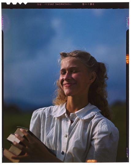 A young woman smiles as she holds a stack of books in her hands. She wears a white blouse with rolled-up sleeves and appears to be standing outdoors, possibly on a hillside or near a field, against a backdrop that suggests it is dusk or dawn due to the blue sky mixed with darker hues above. The image has been captured using color transparency film, as evidenced by the distinctive colors around the edges of the photograph which indicate its age. This particular photograph was taken in September 1947 for Collier's magazine and forms part of a collection called "New England Characters."