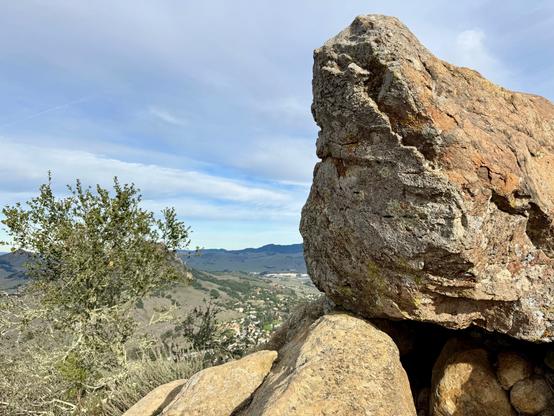 Huge boulders sit on top of one another comprising the summit of Cerro San Luis mountain in San Luis Obispo, California. Behind the top rock is blue sky streaked with clouds. Also in the distance is a shrub, and behind the shrub (hard to make out) is Bishop Peak. Even more mountains are beyond Bishop Peak.