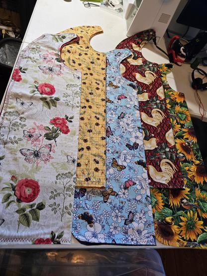 Handmade adult bibs in 100%cotton, bright, bold patterns- left to right-

 red, pink and mauve roses and butterflies, with leaves in shades of green, black screenprint outlines on white,

 bees, flowers, berries and cedar branches against yellow/white gingham print,

 colored butterflies with white flowers against sky blue,

 shades of cream colored roosters, green leaves and berries set against dark red, 

rich golden yellow sunflowers and bright green leaves against forest green.