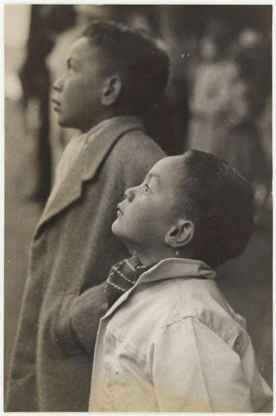 The image is a black and white photograph depicting two young boys. They appear to be outdoors, as suggested by the soft-focus background that hints at an urban environment with indistinct figures of people or objects in motion.

The boy on the left stands taller than his companion, wearing what appears to be a woolen coat typical of mid-20th-century fashion. His posture is upright and attentive, looking off into the distance as if watching something happening out-of-frame.

In contrast, the younger-looking boy on the right has short hair and wears a lighter-colored overcoat with darker trousers or skirt peeking from beneath it. He gazes upward towards his companion's neck, where a small object dangles, which could be a charm or keychain. His expression is one of curiosity or admiration.

Both boys exhibit signs of wear on their coats; the younger boy has noticeable dirt marks on the lower part of his coat, suggesting they have been outdoors for some time and possibly playing outside. The lighting in the photograph highlights the contours of their faces, emphasizing a sense of candidness to this moment captured in time.

The image evokes feelings of nostalgia and innocence, capturing a simple yet poignant childhood memory. Additional context about this particular photo might be found through its citation as being sourced from "Children" by Lyttleton, which could provide historical or biographical details on the subjects  [...]