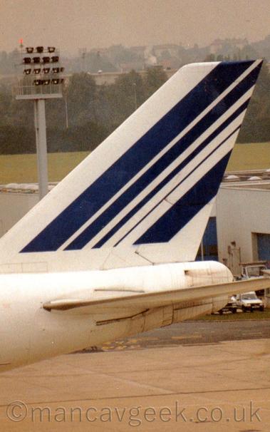 Close-up of the tail  of the very large jet airliner taxiing from right to left.
The plane is almost entirely white, with a series of diagonal blue stripes of varying widths on the white tail.
A grey-brown concrete taxiway fills the bottom of the frame in the background, with a white building with large blue doors beyond that.
Green grass can be seen over the top of the plane, leading up to trees in the distance,, with houses beyond that slowly vanishing into hazy grey sky.