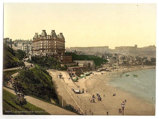 This image depicts a vintage view of the Grand Hotel in Scarborough, Yorkshire, England. The photograph showcases an aerial perspective overlooking the hotel and its surroundings. In the foreground, there is a sandy beach with people scattered about, some walking along the shore while others are seated or standing near green tents. A series of lamp posts line one side of the promenade leading down to the sea.

The Grand Hotel itself stands prominently in the middle ground, an ornate structure with multiple levels and decorative features that exude a sense of grandeur from its design reminiscent of late Victorian architecture. It appears as if it overlooks both the beach area and another hillside dotted with smaller buildings or structures.

In the background, we see more architectural elements including larger edifices possibly representing other historical landmarks in the vicinity, along with hilly terrain that suggests a coastal setting. The overall scene is bathed in warm hues suggesting either sunrise or sunset lighting conditions typical of early photographic processes like photochrom prints from around 1890 to 1900.

This image offers insight into how Scarborough and its Grand Hotel might have appeared during the late Victorian era, highlighting not only architectural features but also daily life activities along a seaside promenade.