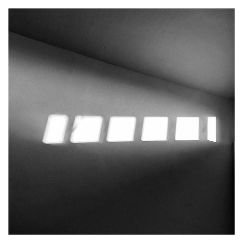Black and white square photo of a row of square windows reflecting sunlight against a wall with a diagonal line above where the wall joins the ceiling
