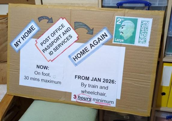 Home-made protest sign. Brown cardboard with a white rectangle where an address label would go. A giant stamp in the top right corner, with the King's face modified with a black Sharpie to look angry. Text on sign: My home > Post Office passport and ID services > Home again. Now: On foot, 30 mins maximum. From Jan 2026: by train and wheelchair, 3 hours minimum.