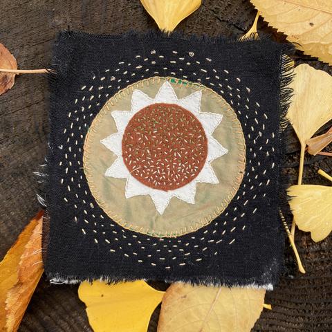 A square of black linen with a circle of gold silk surrounding a 12 pointed white star,  which surrounds a circle of rust colored wool, embellished with simple hand stitching.