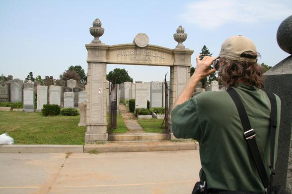 photo looking over the shoulder of a longhaired person in a baseball cap who is himself taking a photo with a camera. the view is looking across a road at a gated cemetery section. at the top of the gate is a harp logo and the words PROG. MUSICAL BEN. ASS'N.