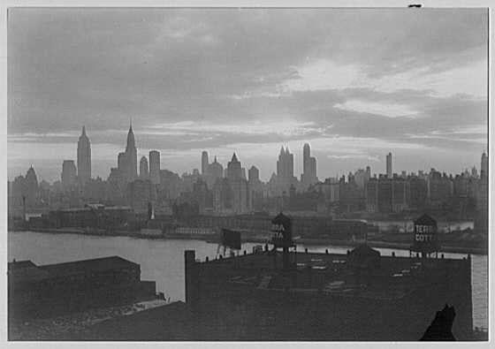 The image is a black and white photograph depicting an urban skyline, presumably of New York City. The scene shows tall skyscrapers in the background with various architectural styles indicative of early 20th-century construction, such as Art Deco designs. Notable buildings like the Empire State Building are visible among others.

In the foreground, there is a body of water that could be part of the East River or Hudson River, which meanders through Manhattan. The skyline appears to overlook this river with numerous industrial structures and rooftops at lower elevations in the immediate vicinity, including what seems to be old warehouses or manufacturing facilities bearing signs like "Santa Fe" and "Gottlieb Co."

The sky is filled with clouds, suggesting an overcast day or possibly early evening light. The overall atmosphere of the photograph conveys a sense of urban life during that period.

This image is part of a collection titled 'New York City views,' which includes various photographs depicting different aspects of New York from around 1932 to the mid-20th century. Samuel H. Gottscho, whose name appears alongside this image, was known for his work as an architectural photographer and real estate developer in New York during that era.

The reference provided indicates a specific date—April 12, 1932—which might be relevant to the context or significance of the photograph within its collection. The negative dimension [...]