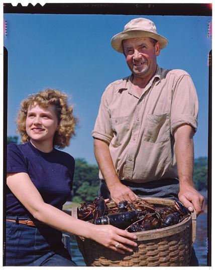 The image depicts two individuals outdoors on a sunny day. The person on the left is smiling, with curly blonde hair and wearing a dark blue t-shirt paired with denim jeans. They are holding onto a basket containing lobsters or crabs.

The individual on the right appears to be an older man wearing a beige hat, light-colored long-sleeve shirt, khaki pants, and sunglasses pushed up towards his forehead. He is standing behind the basket of crustaceans, also smiling at the camera with hands resting lightly on its edge.

Both individuals are seated beside what seems like water or a boat, suggesting they may have been involved in fishing activities. The background shows trees under clear blue skies, indicating pleasant weather conditions for outdoor activities.