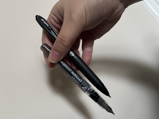my hand holding two black pens. a click uniball pen that is narrow with a sleek soft grip and a very small hook. a semi-transparent and thicker casing for the fountain pen.