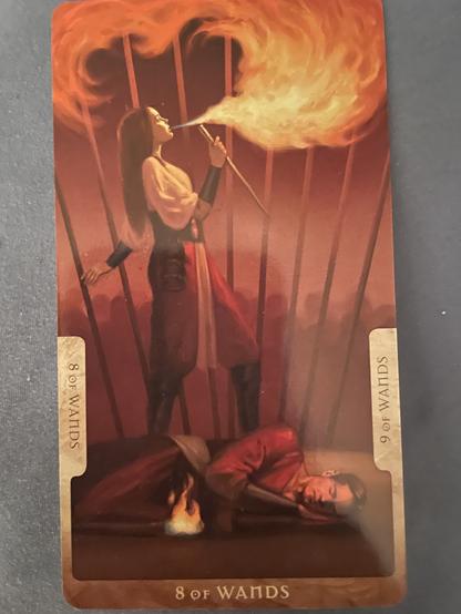 A man in red lies on the ground, resting. A woman behind him blows fire over a wand she holds. A further line of wands separates them both from a faceless crowd opposing them.