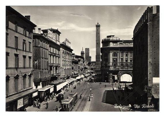 The black and white photograph depicts a bustling urban scene with vintage architecture. The image is labeled "Bologna - Via Rizzoli," indicating the location as via Rizzoli in Bologna, Italy. Tall buildings line both sides of a busy street filled with pedestrians, tram cars, and horse-drawn carriages, showcasing early 20th-century transportation methods.

The central figure in the photograph is the Torri Campeggi (also known as Palazzo d'Accursio), a historic tower that stands prominently amidst the cityscape. Surrounding it are various commercial establishments with signs such as "Campari" and others advertising products like beverages, suggesting an active marketplace.

Further down the street, more historical buildings can be seen, including one with arched windows and another featuring ornate detailing at its top edge. The presence of these structures suggests a rich architectural history in Bologna's urban fabric.

The overall scene captures daily life in early 20th-century Italy as it unfolds on this particular via Rizzoli street, offering a glimpse into the city's past before modern advancements transformed public spaces and transportation systems.
