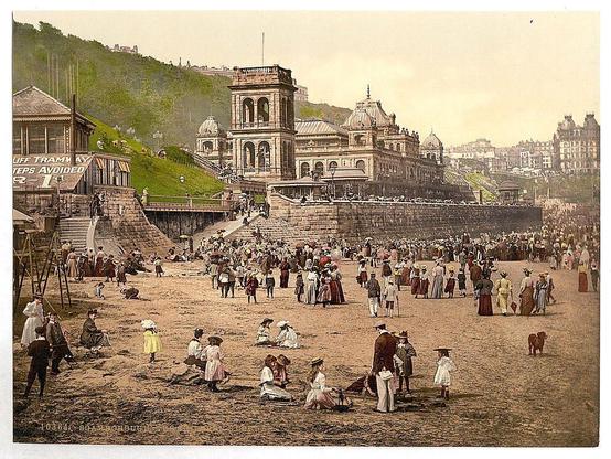 The image depicts a bustling beach scene at the Spa in Scarborough, North Yorkshire, England. The photograph captures an early 20th-century setting with numerous people milling about on the sandy shore and ascending steps leading up to grand buildings indicative of a spa complex. Individuals are dressed in period-appropriate attire, including hats and long dresses for women and suits for men, suggesting leisurely social activities typical of Victorian-era seaside resorts.

The Spa's architecture features ornate facades with domed roofs and prominent signage indicating its purpose as an entertainment venue or health facility. The backdrop includes a hilly landscape adorned with residential buildings and greenery, contributing to the picturesque quality of the scene. Several people are engaged in various activities such as walking along the shore, sitting on steps, conversing in groups, and observing their surroundings.

The photograph has aged tones typical of early 20th-century colorized images, giving it a historical ambiance that transports viewers back to an era when seaside resorts were popular destinations for relaxation and social gatherings.