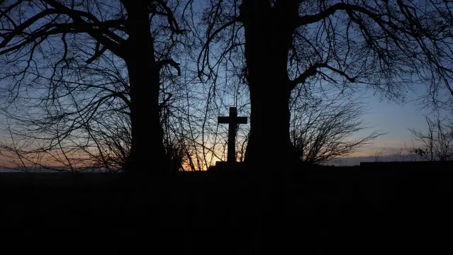 Zwischen zwei Bäumen steht ein Kreuz, alles als Schattenriss vor einem dunkelblauen Himmel.