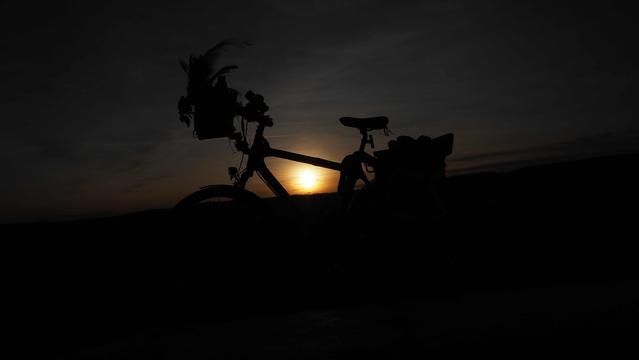 Schattenriss eines Fahrrades mit Lenkerkorb vor dem Sonnenball der durch den Rahmen des Rades scheint. Drumherum ein dunkelgrauer Himmel