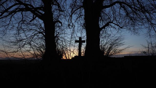 Zwischen zwei Bäumen steht ein Kreuz, alles als Schattenriss vor einem dunkelblauen Himmel.