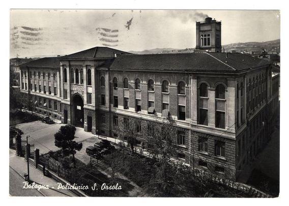 The image depicts a historic building labeled "Bologna - Policlínico S. Orsola," which appears to be an old photograph, possibly dating back several decades given its black and white quality and the style of cars visible in front of it. The structure is multi-storied with symmetrical design elements including arched windows and a tower at one end. It has classical architectural features such as tall columns supporting arches above large doorways. There are trees around, indicative that this building could be situated on or near an urban park.

The photograph shows the entrance of the polyclinic which is marked by a prominent signboard stating "Policlinico S." suggesting it's part of a larger medical facility network in Bologna, Italy. The surrounding environment includes other buildings and trees with some leafless branches indicating that the season might be late autumn or winter.

The sky is overcast, providing muted natural lighting which contributes to the vintage feel of the photograph. There are no visible people within this particular shot, focusing attention on the architecture itself. The image conveys a sense of historical significance and urban development in Bologna during its time.