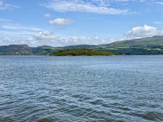 A calm expanse of water stretches across the foreground, with gentle ripples reflecting the daylight under a partly cloudy sky. Beyond the water lies a small green island with low vegetation, bordered by rocky edges. Rolling hills rise behind it, covered in a mix of grassland and patches of woodland, with some areas appearing darker where the land meets shadow. Further in the distance, taller hills and ridges form a layered backdrop, their slopes varying in shades of green and brown. White clouds drift across the bright blue sky, adding contrast to the landscape. The overall scene is quiet and open, with no visible human activity on the water.