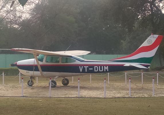 A small aircraft with the registration VT-DUM is displayed outdoors, surrounded by a low fence. It features the Indian flag colors on its tail.