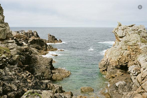 Felsküste mit zerklüfteten Klippen und einem schmalen Meeresarm, in dem grün-blaues Wasser zwischen den Felsen in den offenen, leicht bewölkten Ozean hinausfließt.